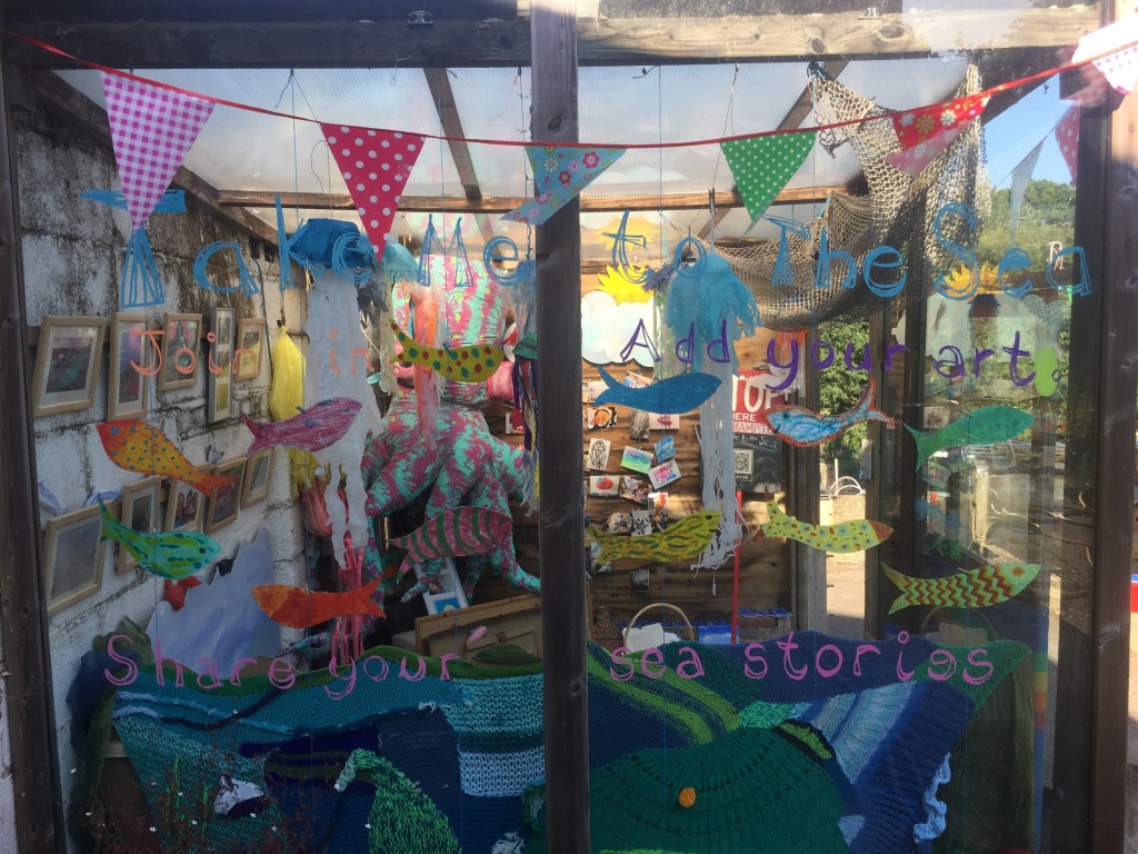 A picture of a glass house full of arty thing.s, including hand painted fish, knitted waves, postcards and framed pictures, a large octopus and paper jellyfish.